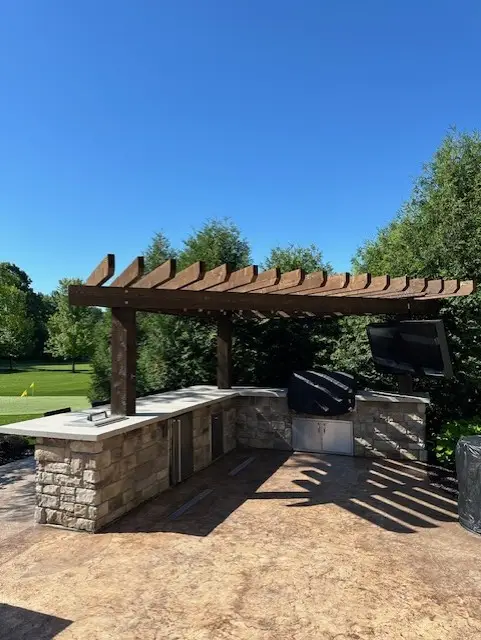 "Elegant backyard kitchen in Hartland, WI, showcasing a natural stone L-shaped bar with concrete countertops, a grille, and a refrigerator. Complete with a putting green, this setup blends style and function for a perfect outdoor retreat. #HartlandWI #BackyardKitchen #NaturalStone #ConcreteCountertops #PuttingGreen"