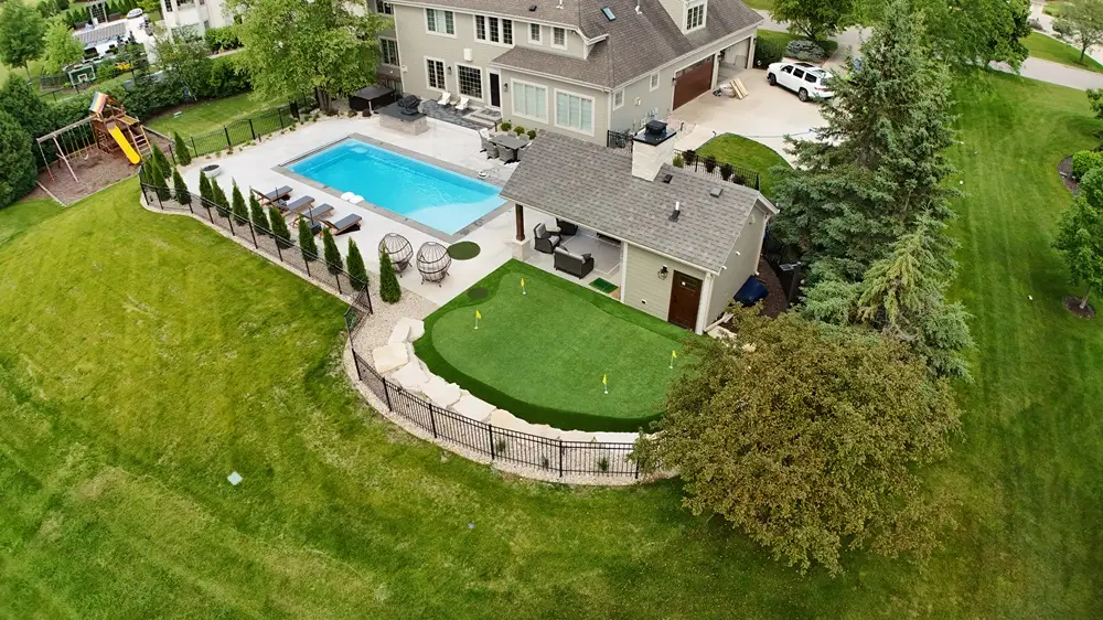 "Transformed outdoor living space featuring a luxurious pool, lush plantings, a fenced-in yard, striking stone walls, a versatile outdoor structure, and a fun putting green. This beautifully designed area combines relaxation, play, and privacy for the ultimate backyard retreat. #OutdoorLiving #PoolDesign #StoneWalls #PuttingGreen #BackyardParadise"